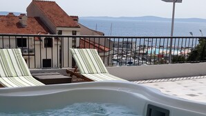 Un jacuzzi con dos sillones reclinables a rayas, vista al mar y un edificio con tejado rojo al fondo.