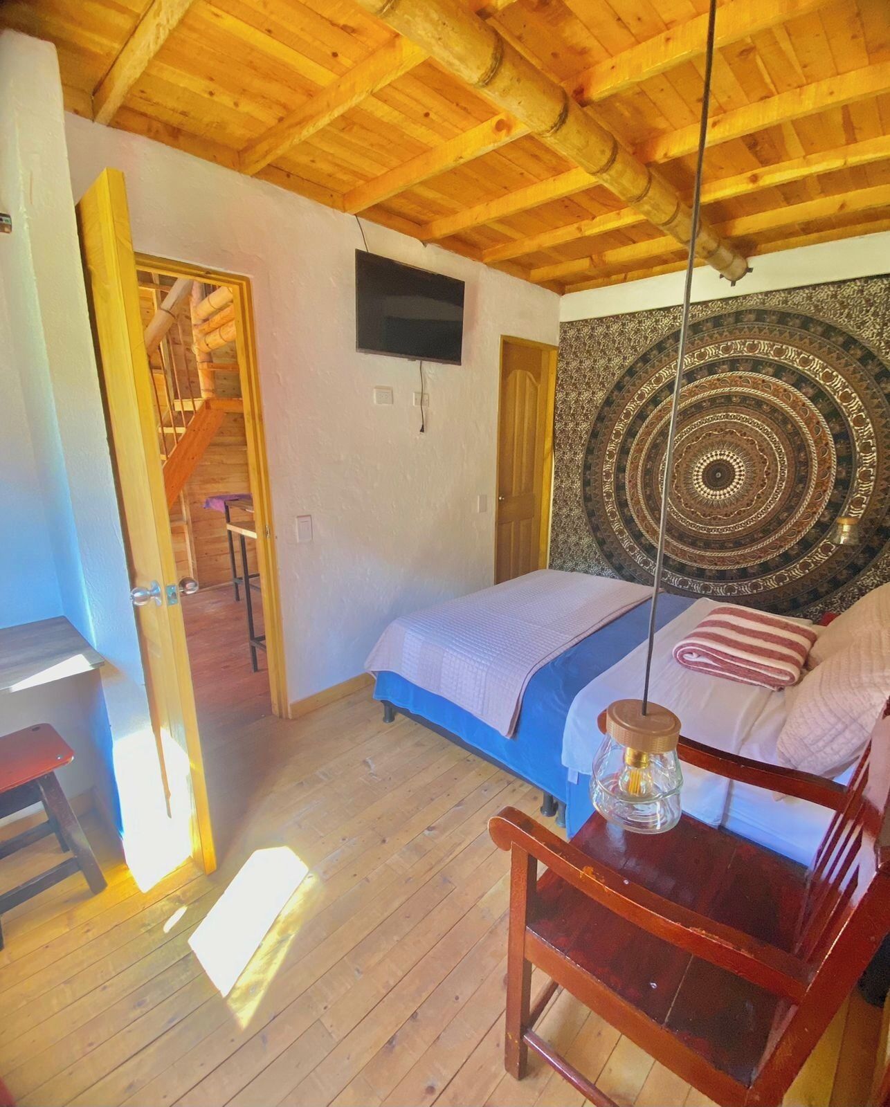 A bedroom with a wooden ceiling, a bed with a blue bedspread, a wooden chair, a tapestry on the wall, and a television mounted above the bed.