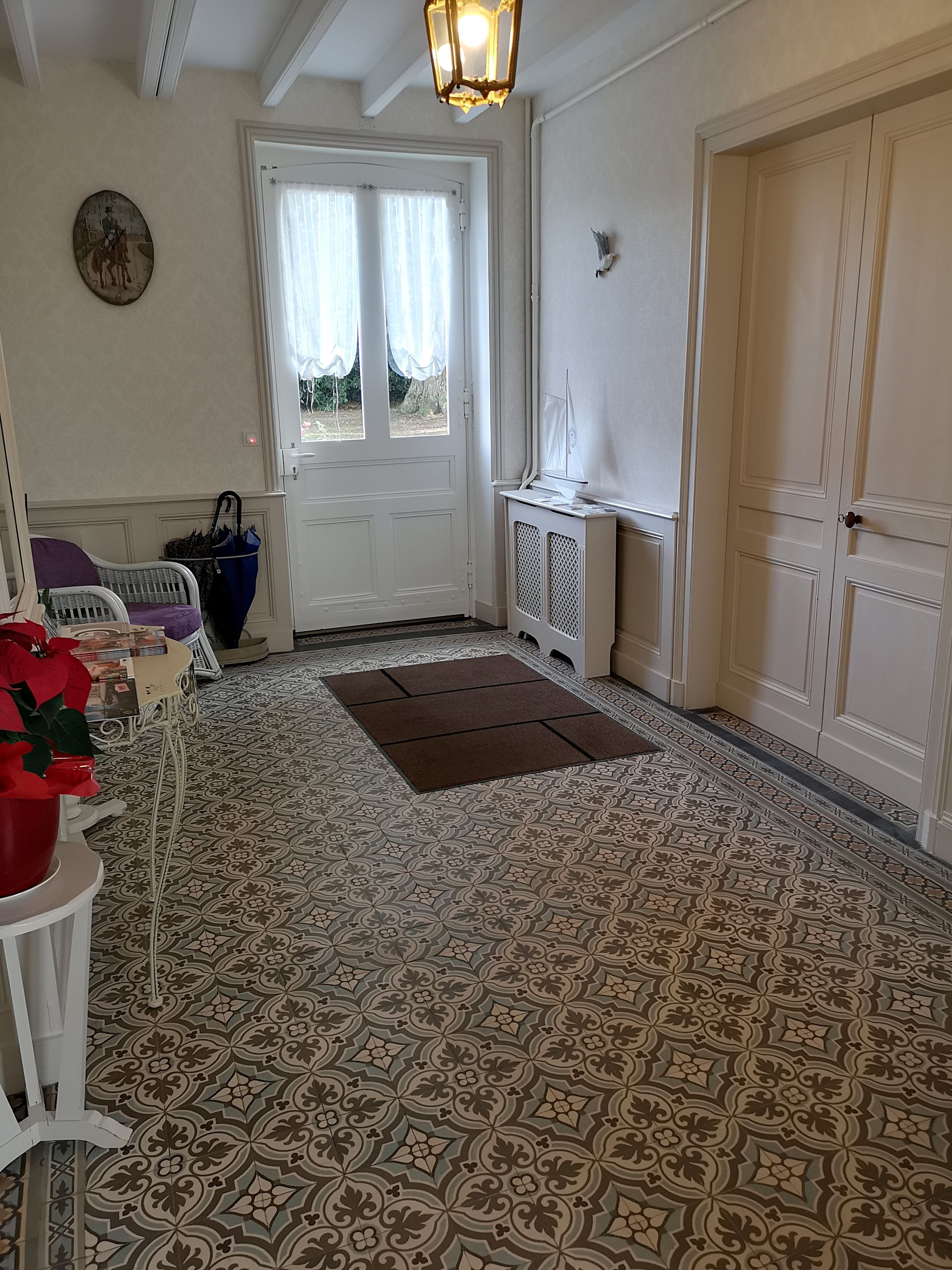 Una habitación con baldosas de suelo estampadas, una puerta blanca con cortinas, una mesa pequeña con una olla roja y una silla blanca.