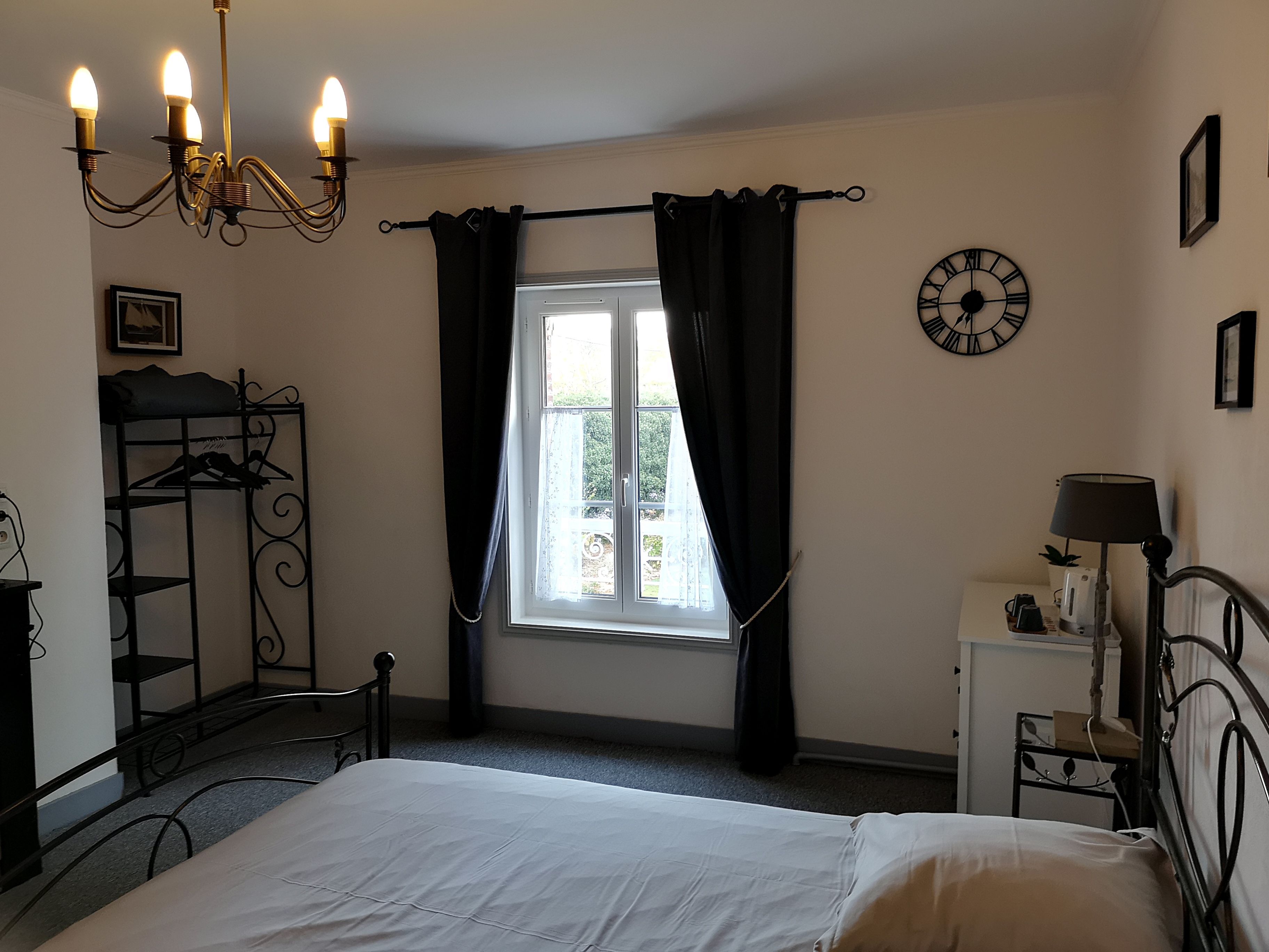 Un dormitorio con cama, ventana con cortinas, un reloj de pared, una mesita de noche con lámpara y un armario metálico.