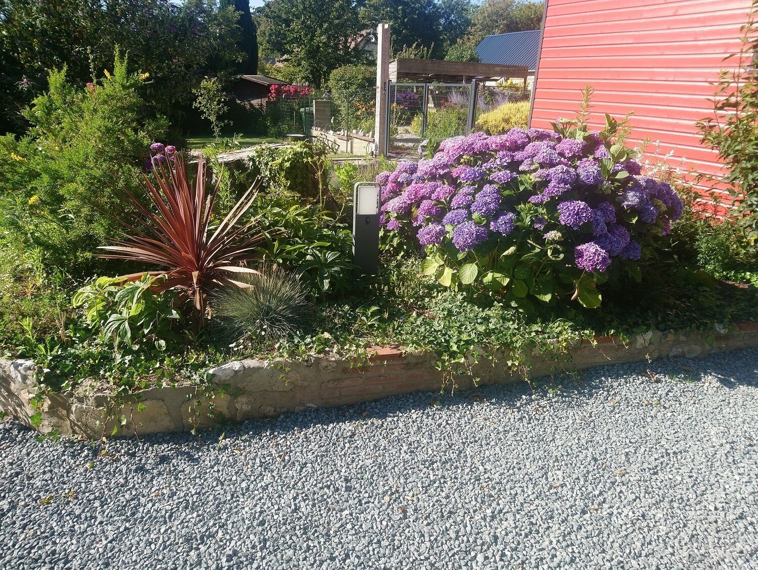 Ein Garten mit verschiedenen Pflanzen und Blumen, einem Kiesweg und einem roten Gebäude im Hintergrund.