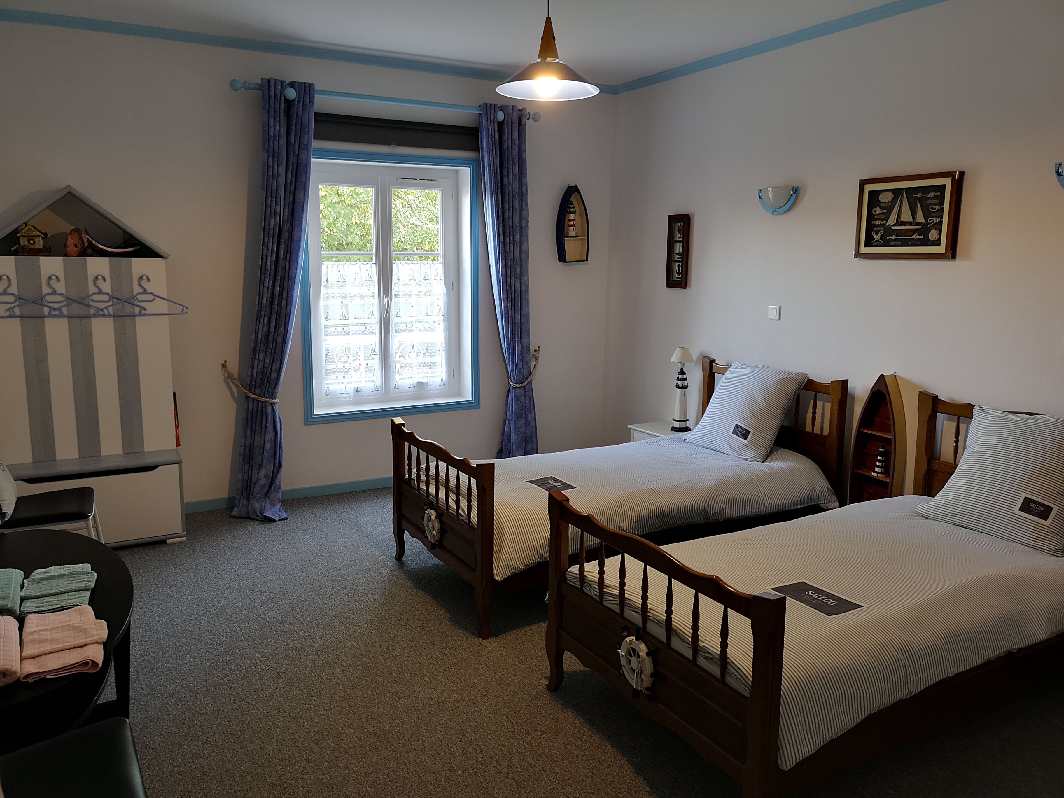 Una habitación con dos camas individuales, una mesita, una silla, un ventanal con cortinas y una lámpara.