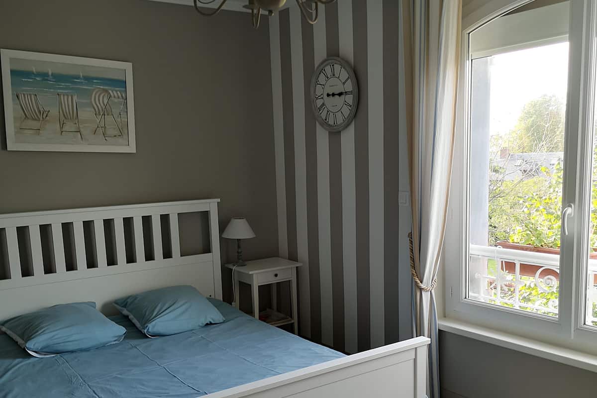 A bedroom with a white bed, a clock on the wall, a window with curtains, and a painting of beach chairs.