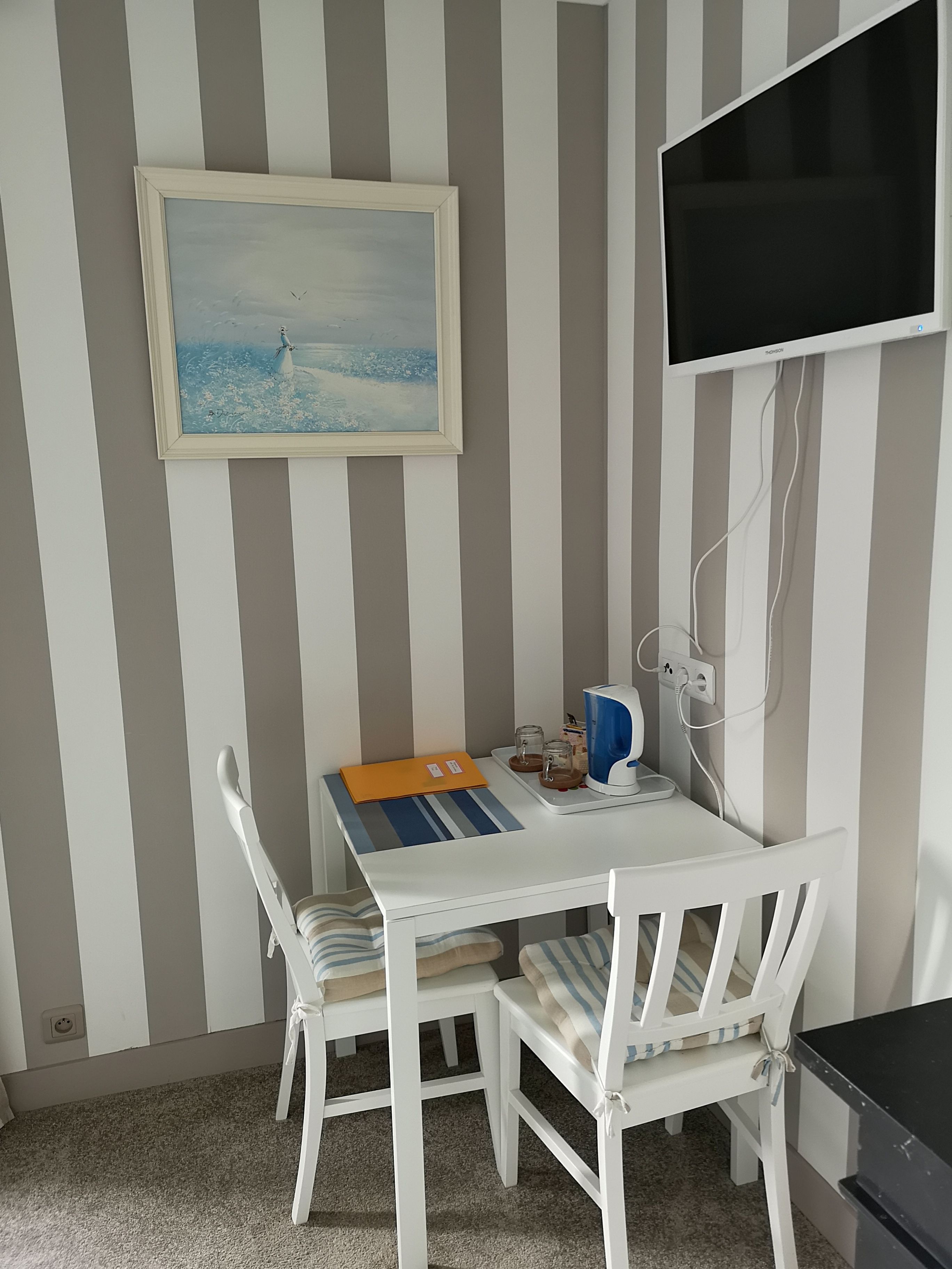 Una habitación con pared a rayas, un cuadro, un televisor, un escritorio con una silla y una mesita con una bandeja.