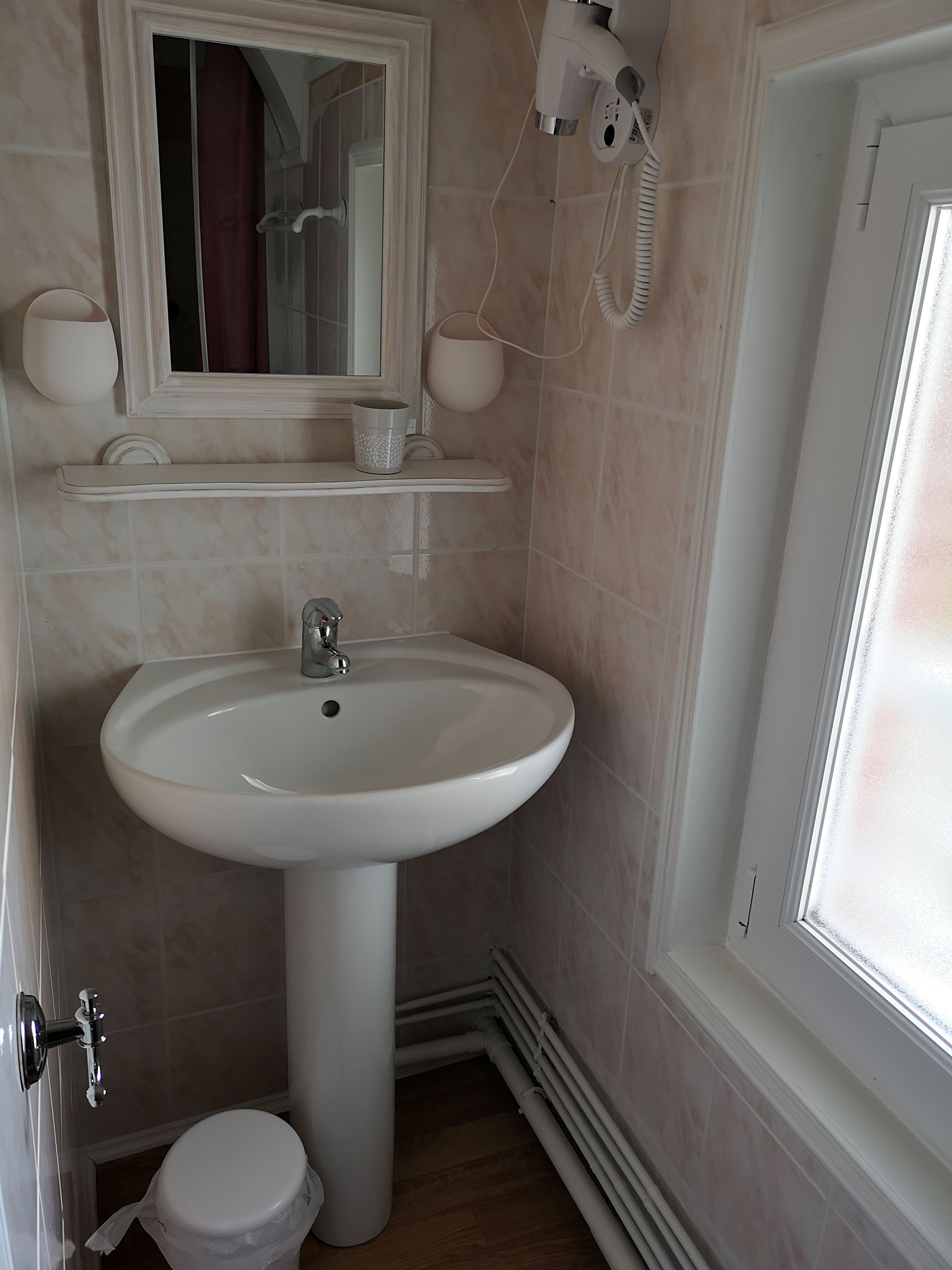 A bathroom with a white pedestal sink, a toilet, a mirror, and a hairdryer mounted on the wall.