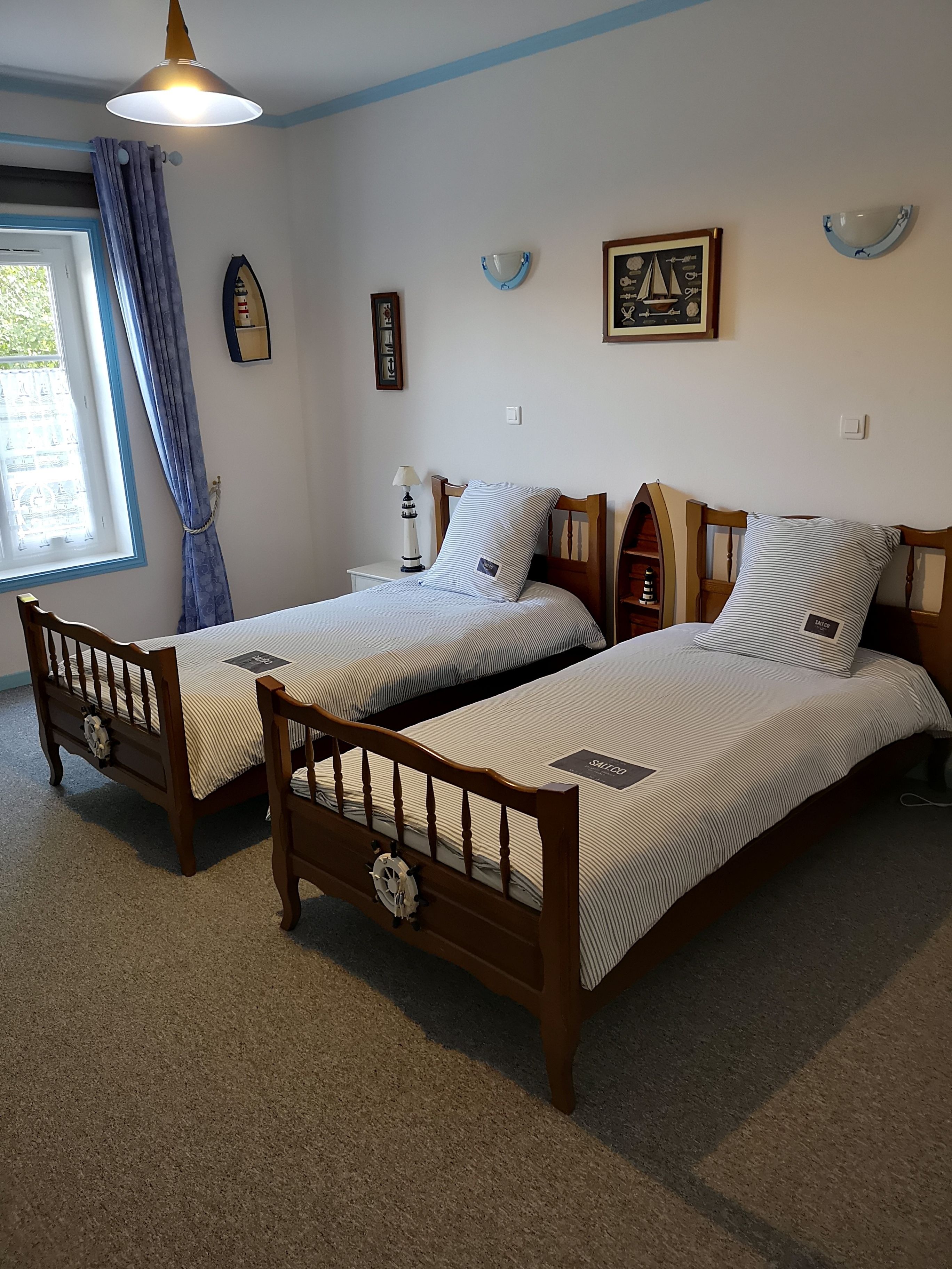 A room with two wooden beds, a window with curtains, a wall-mounted lamp, and a framed picture.