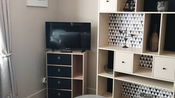 A compact living space with a television on a stand, a small round stool, a patterned floor, and a wall-mounted shelf with decorative items.