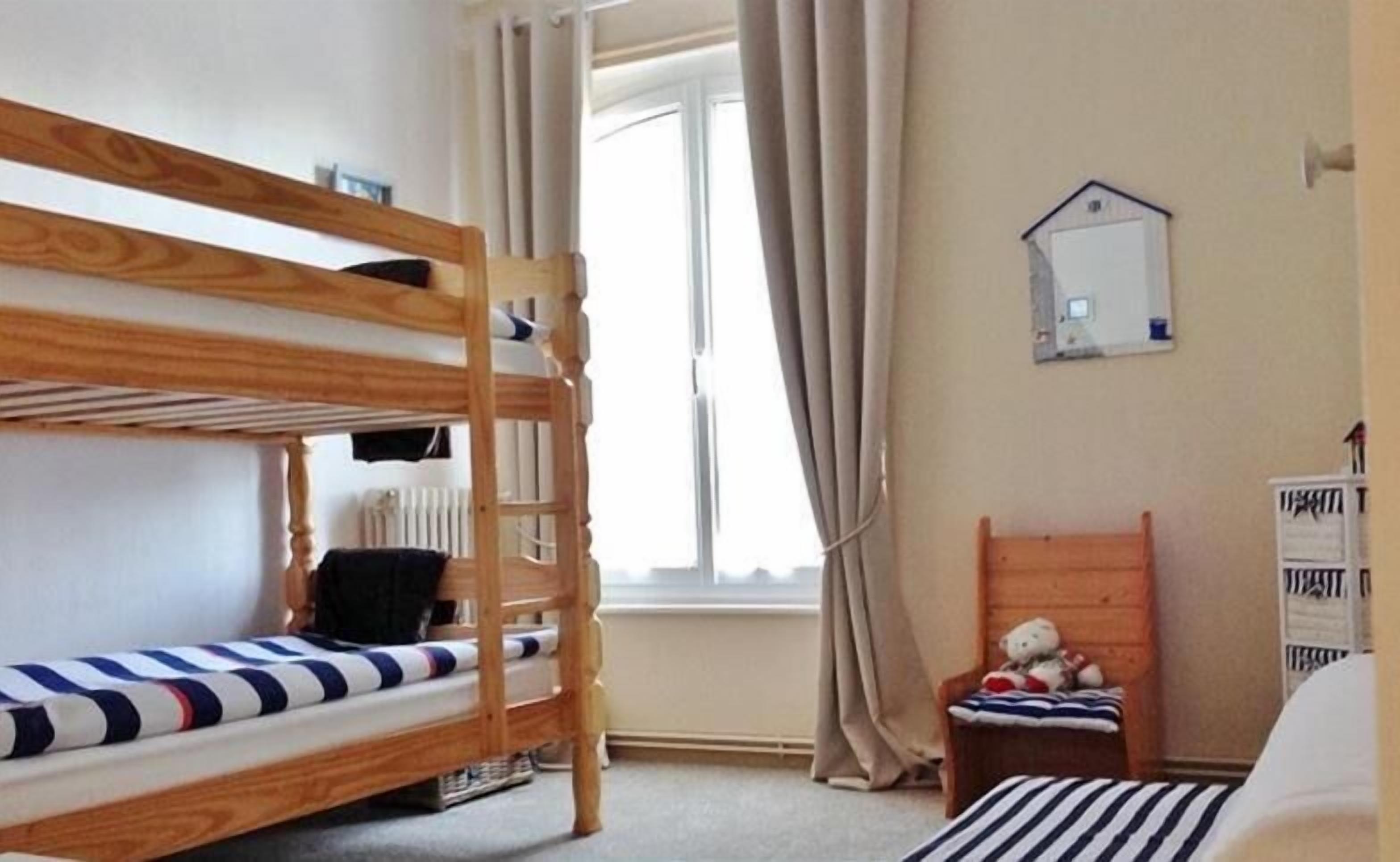 Une chambre avec un lit superposé, une fenêtre, des rideaux, une petite chaise avec un ours en peluche et un miroir au mur.