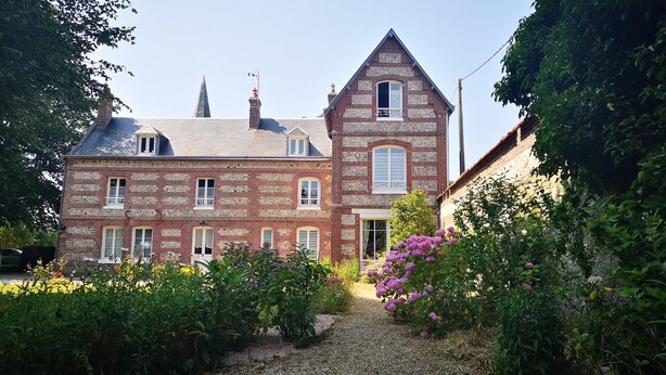 A large, historic brick house with a prominent tower, surrounded by lush gardens and a gravel path leading to the entrance.