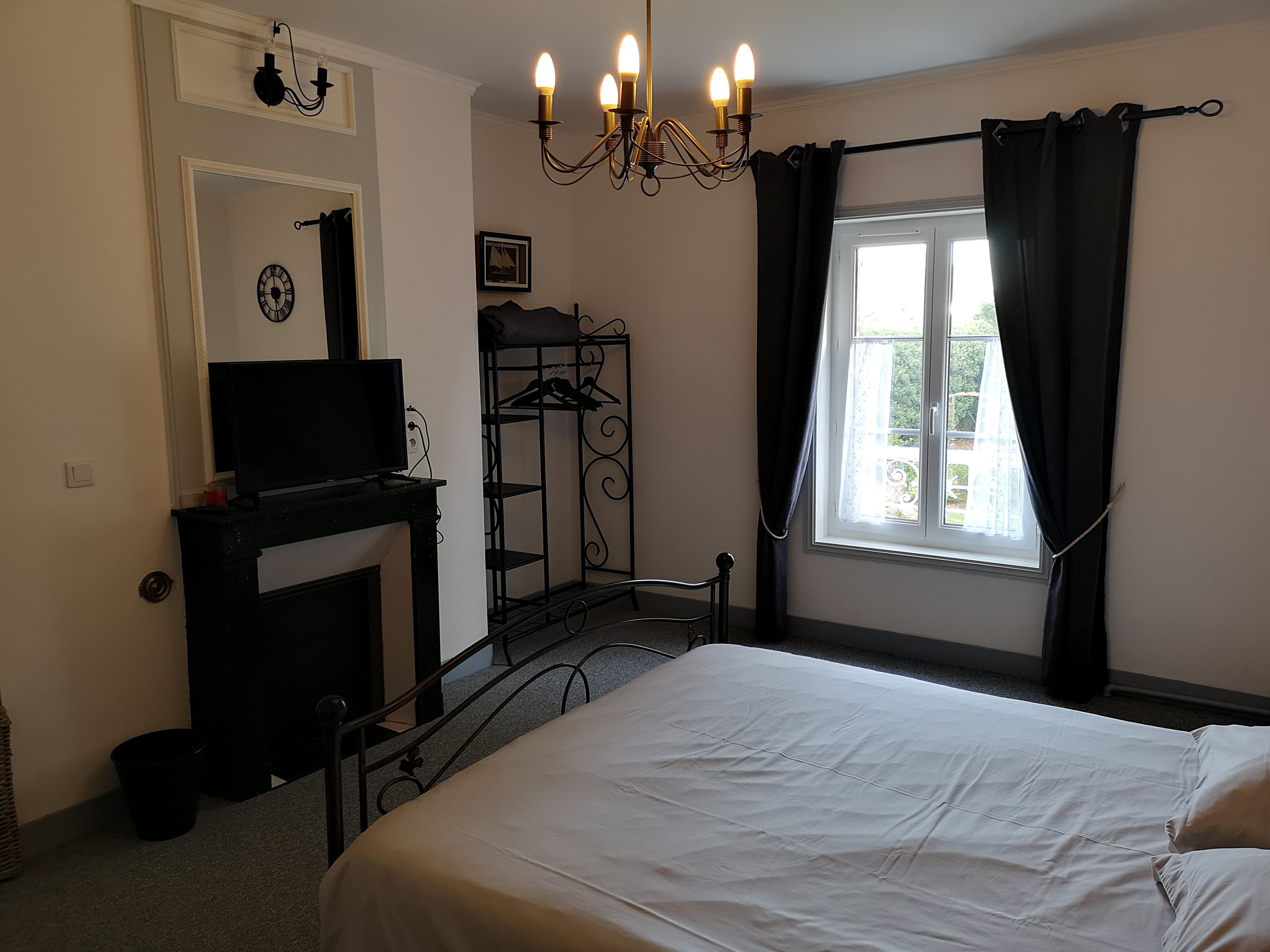 A bedroom with a bed, a television on a dresser, a chandelier, a window with curtains, and a coat rack.