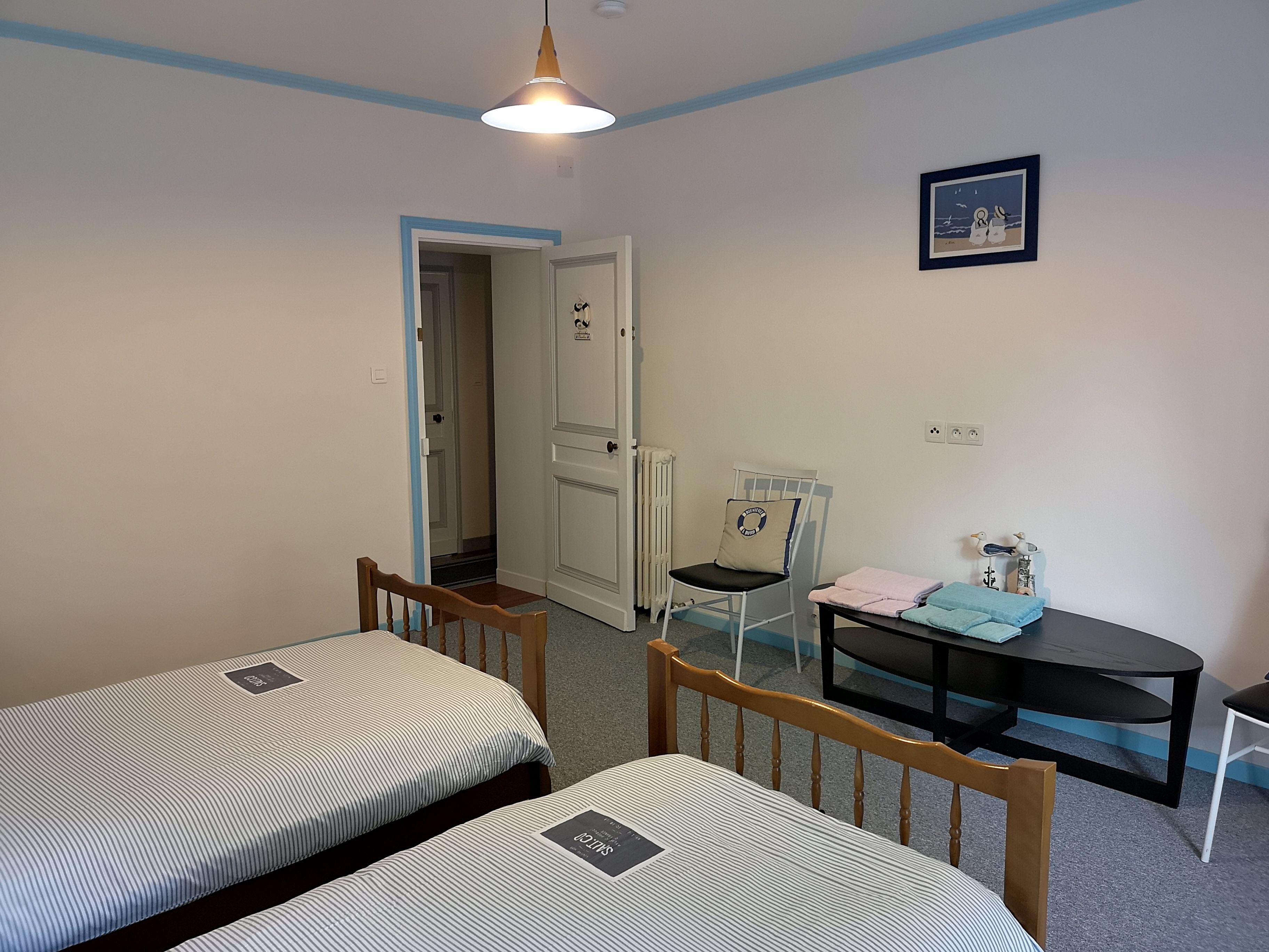 Una habitació amb dos llits individuals, una taula petita amb cadires, un radiador i una porta que condueix a una altra habitació.