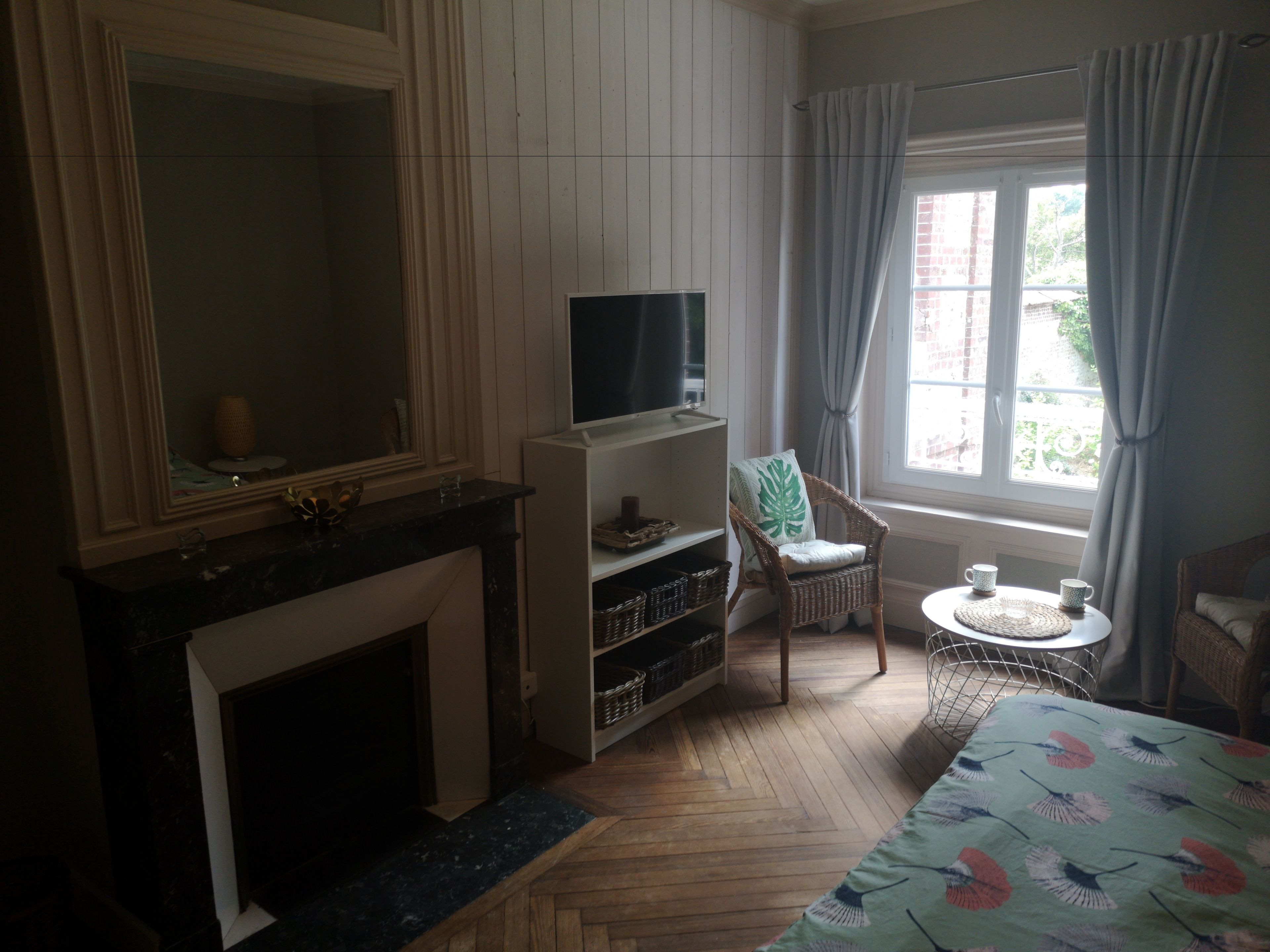 A hotel room with a fireplace, a television, a wicker chair, a small round table with two cups, and a window with curtains.