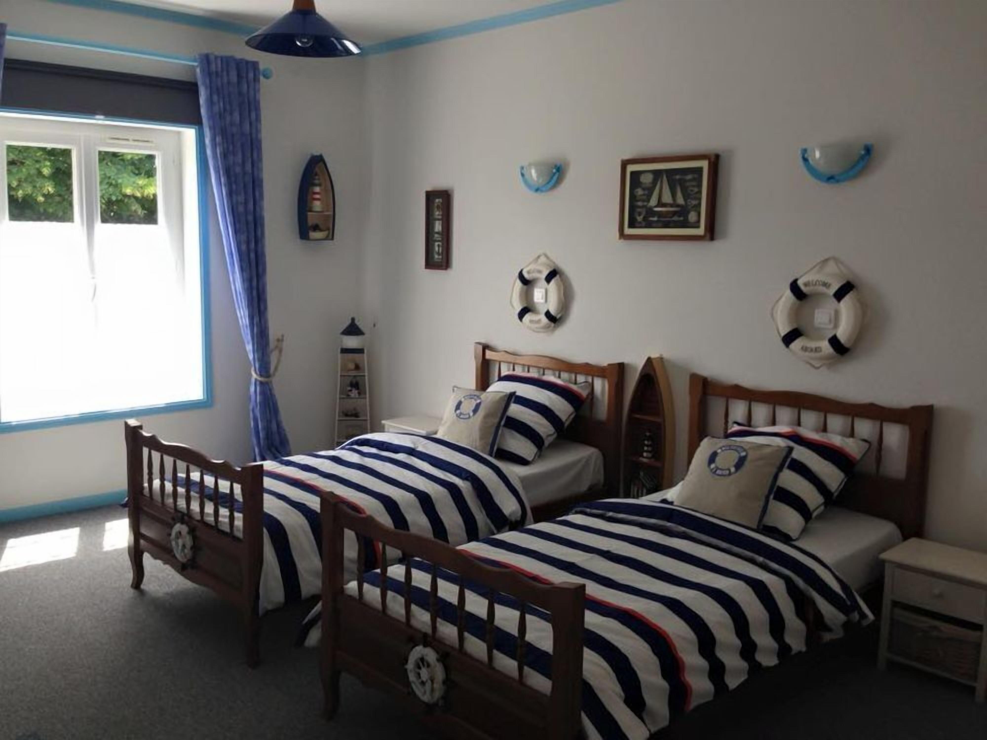 Quarto com duas camas de madeira, uma janela com cortinas azuis e decoração temática náutica.
