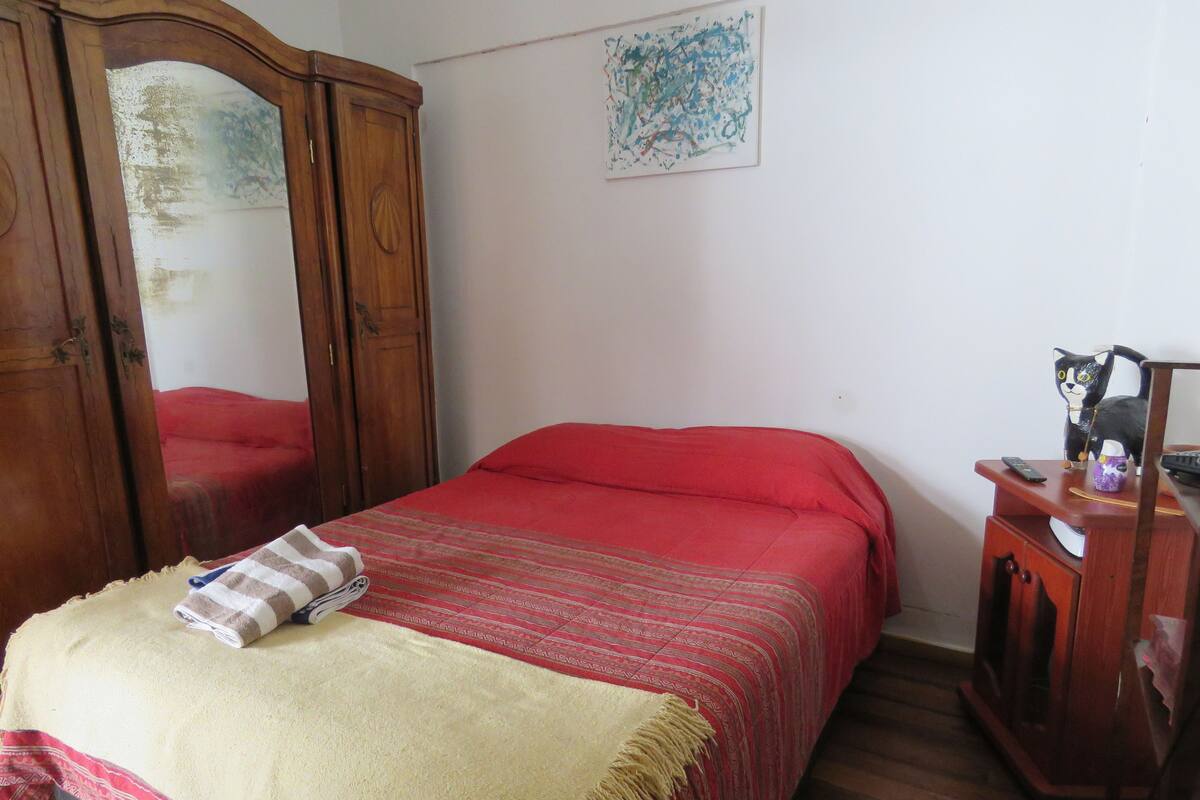 Um quarto com cama, guarda-roupa de madeira, espelho, quadro e uma pequena mesa com uma estatueta.