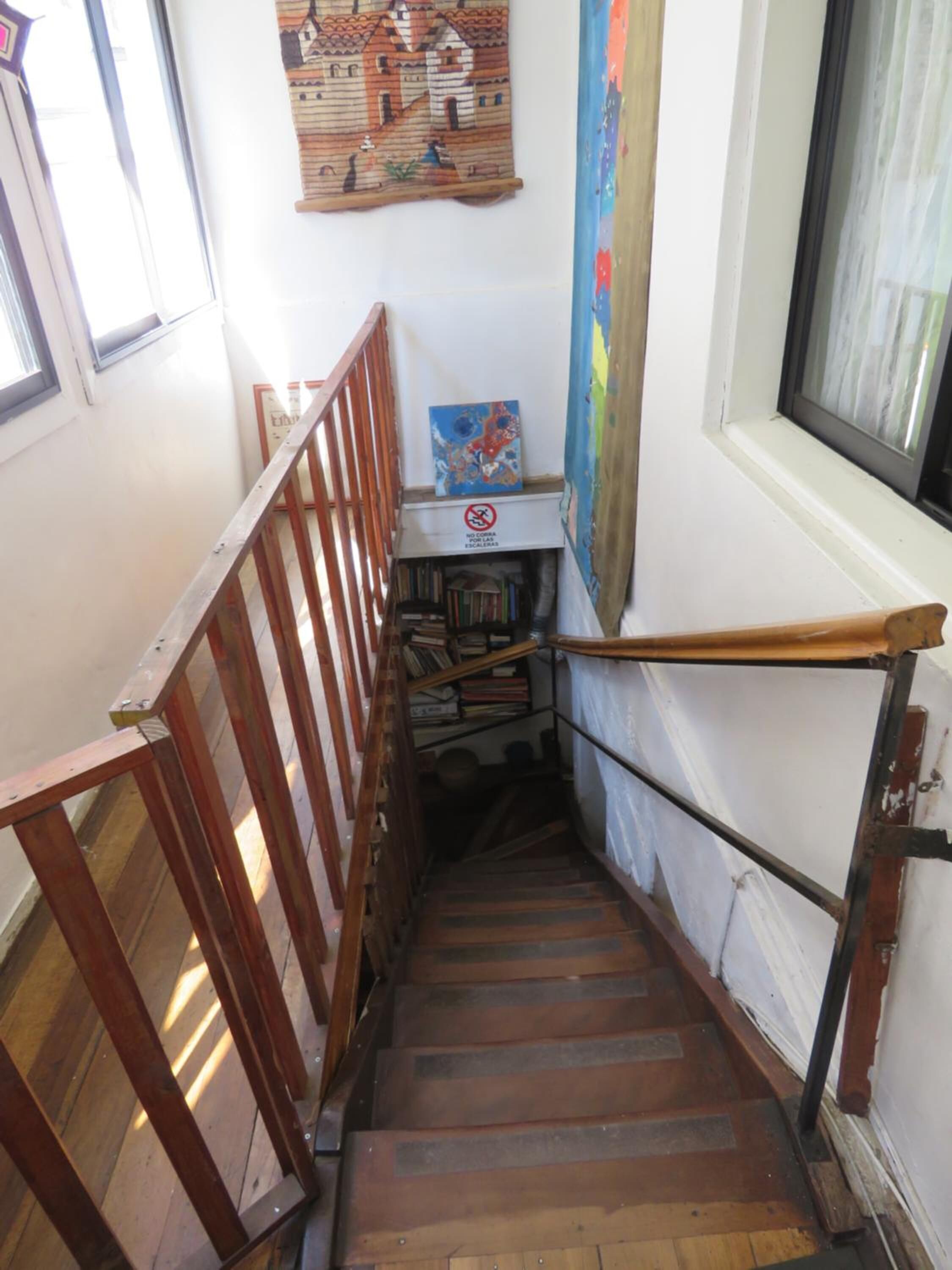 Un escalier étroit en bois, muni d’une main courante, mène à un palier où se trouvent une bibliothèque et un tableau.