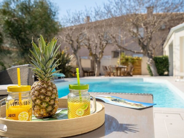 A tray with a pineapple, a glass of fresh juice, and a smoothie with a straw, placed by a pool with a view of a house and trees.