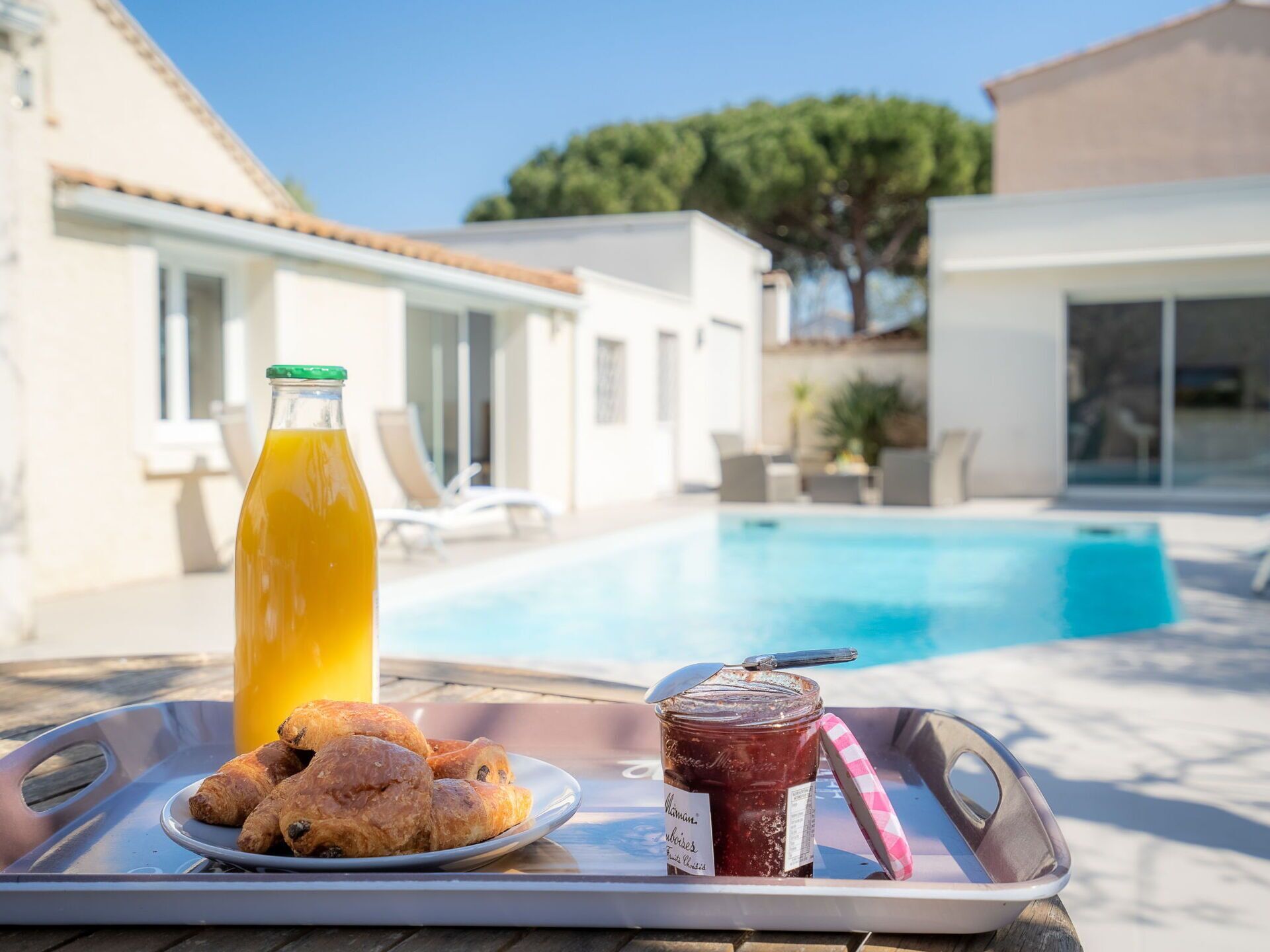 A tray with croissants, a bottle of orange juice, and a jar of jam by a poolside.