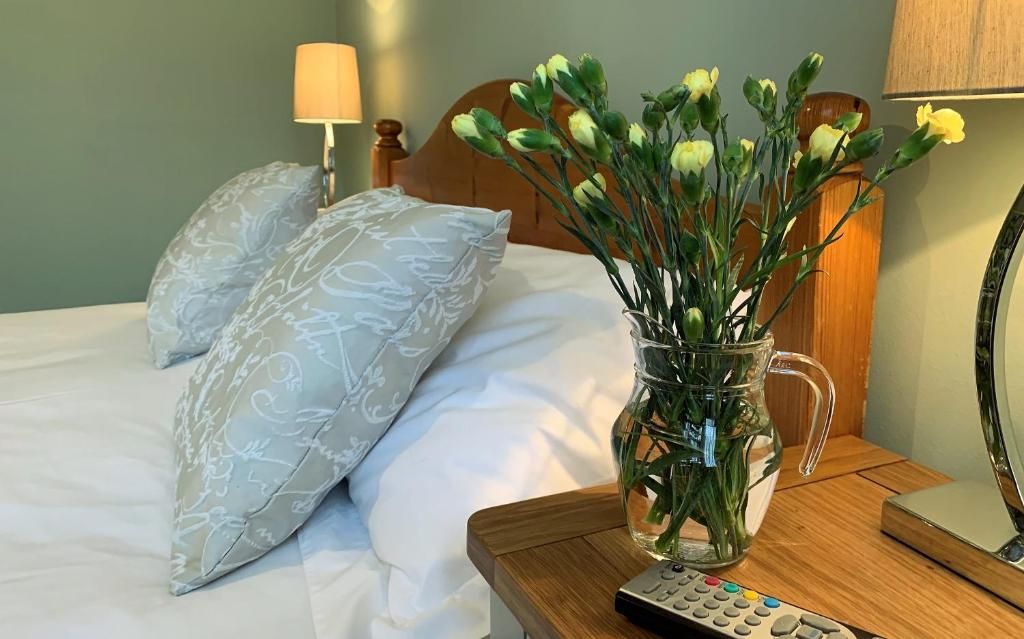 Een bed met witte lakens, een vaas met bloemen, een afstandsbediening en een houten nachtkastje.