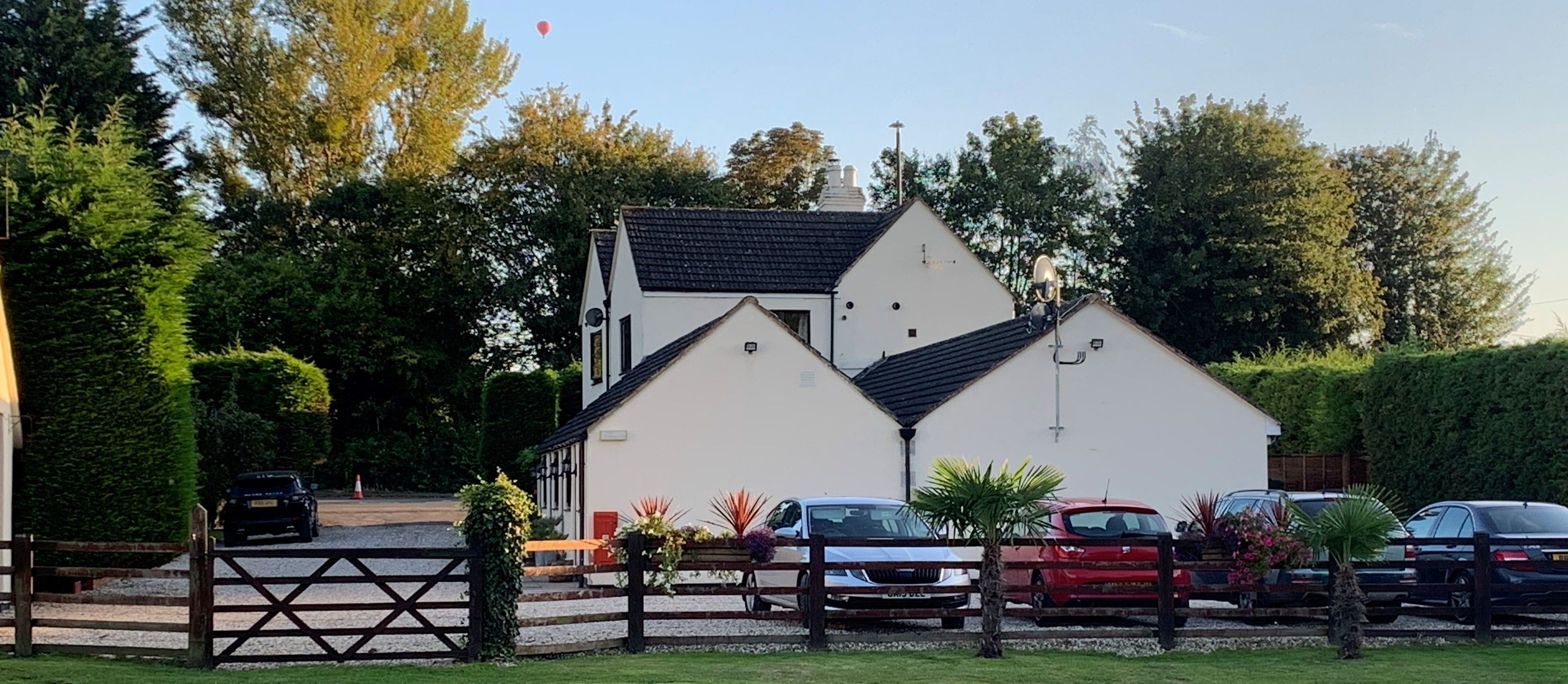 Ein zweistöckiges Gebäude mit weißer Fassade und dunklem Dach, umgeben von parkenden Autos und Grünpflanzen.