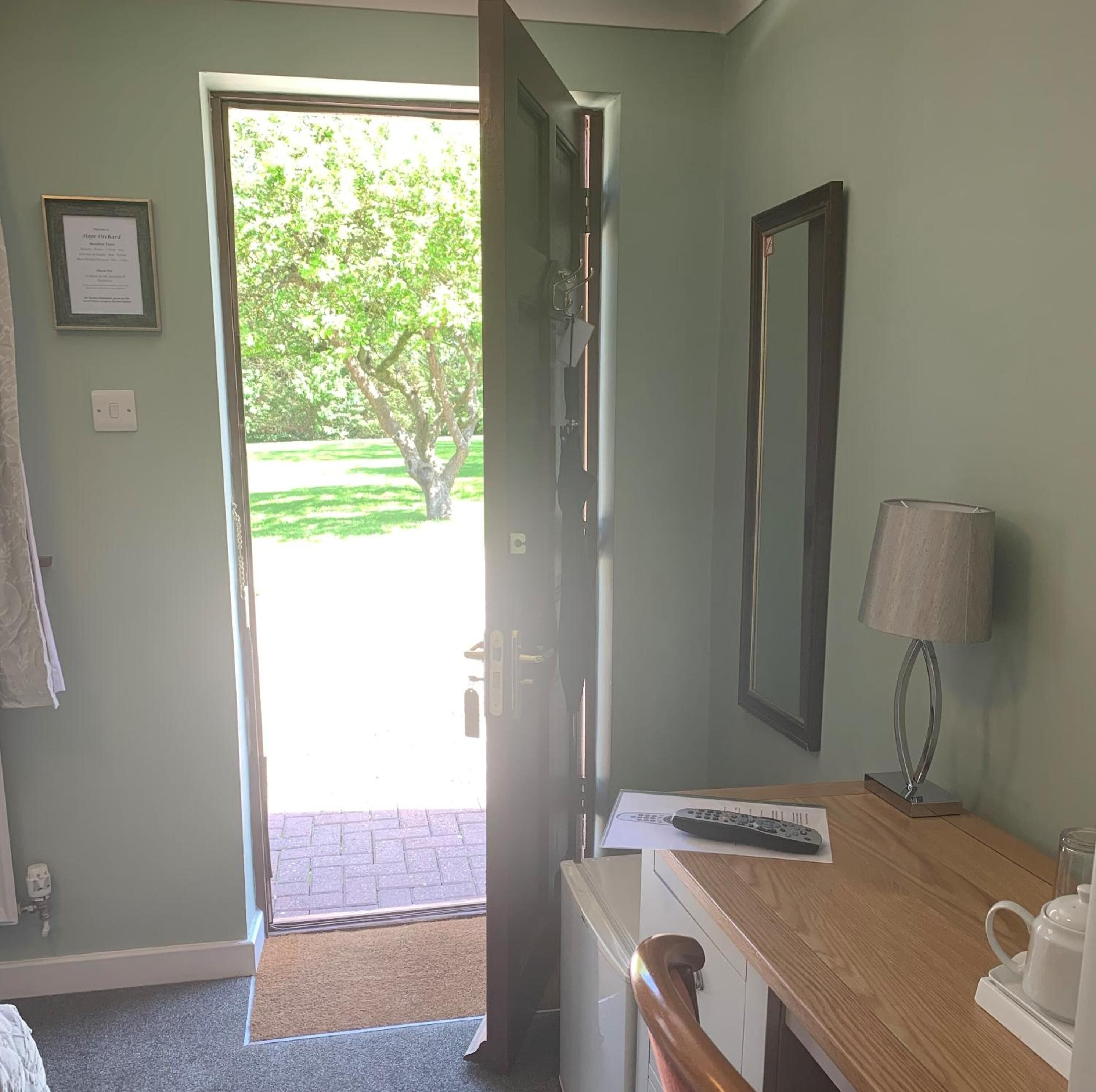 A room with a door leading to a garden, a bedside table with a lamp, a framed certificate on the wall, and a window with a view of trees.