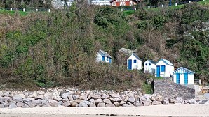 A coastal area with a stone wall, beach, and several small blue and white buildings with steep roofs, likely holiday homes or cottages.