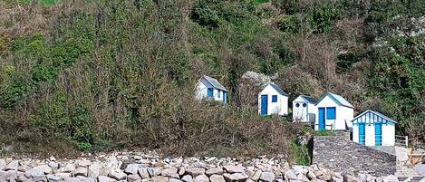 A coastal area with a stone wall, beach, and several small blue and white buildings with steep roofs, likely holiday homes or cottages.