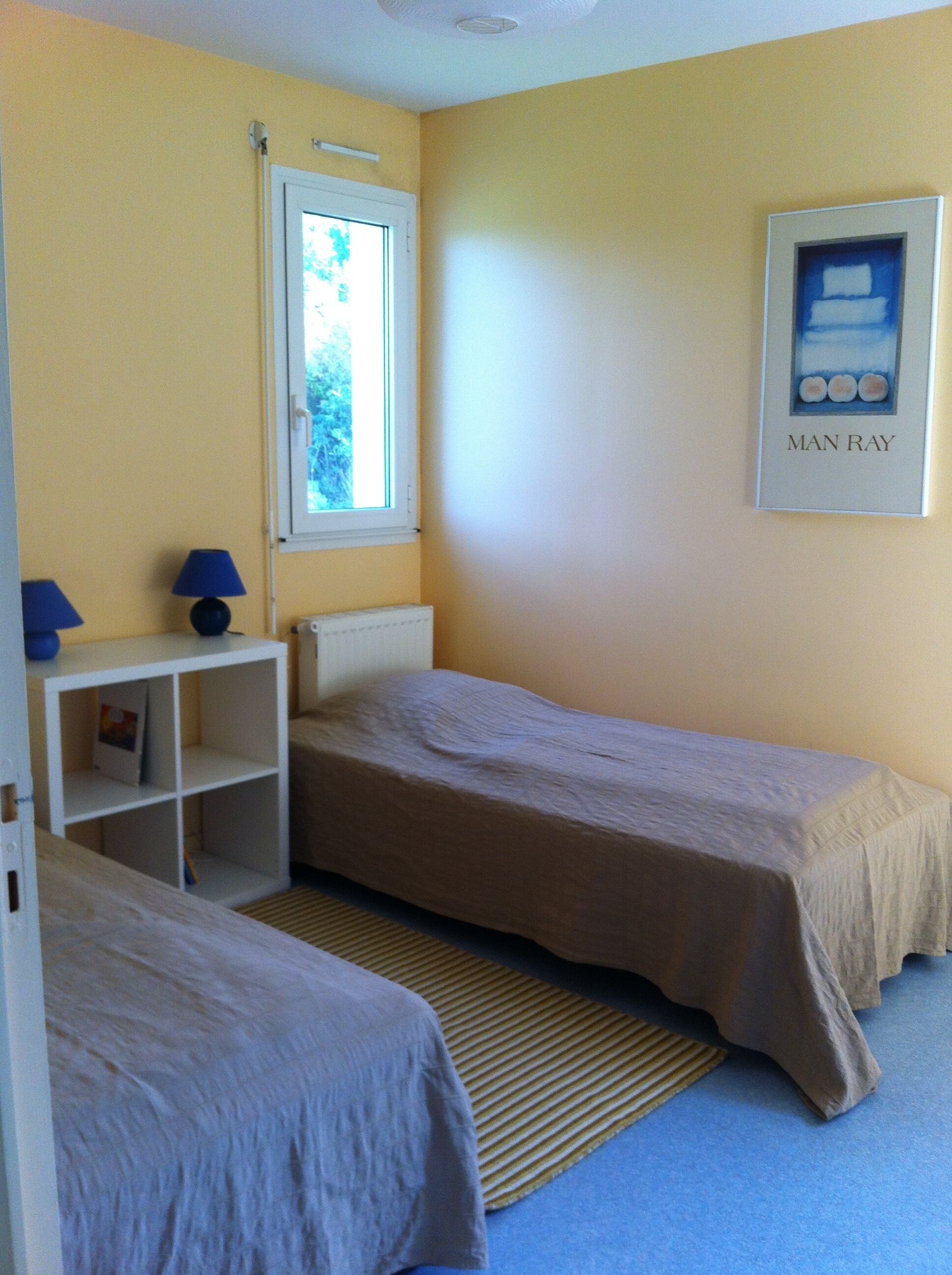 A small, single-room accommodation with two single beds, a window, a shelf with a lamp, and a framed picture on the wall.