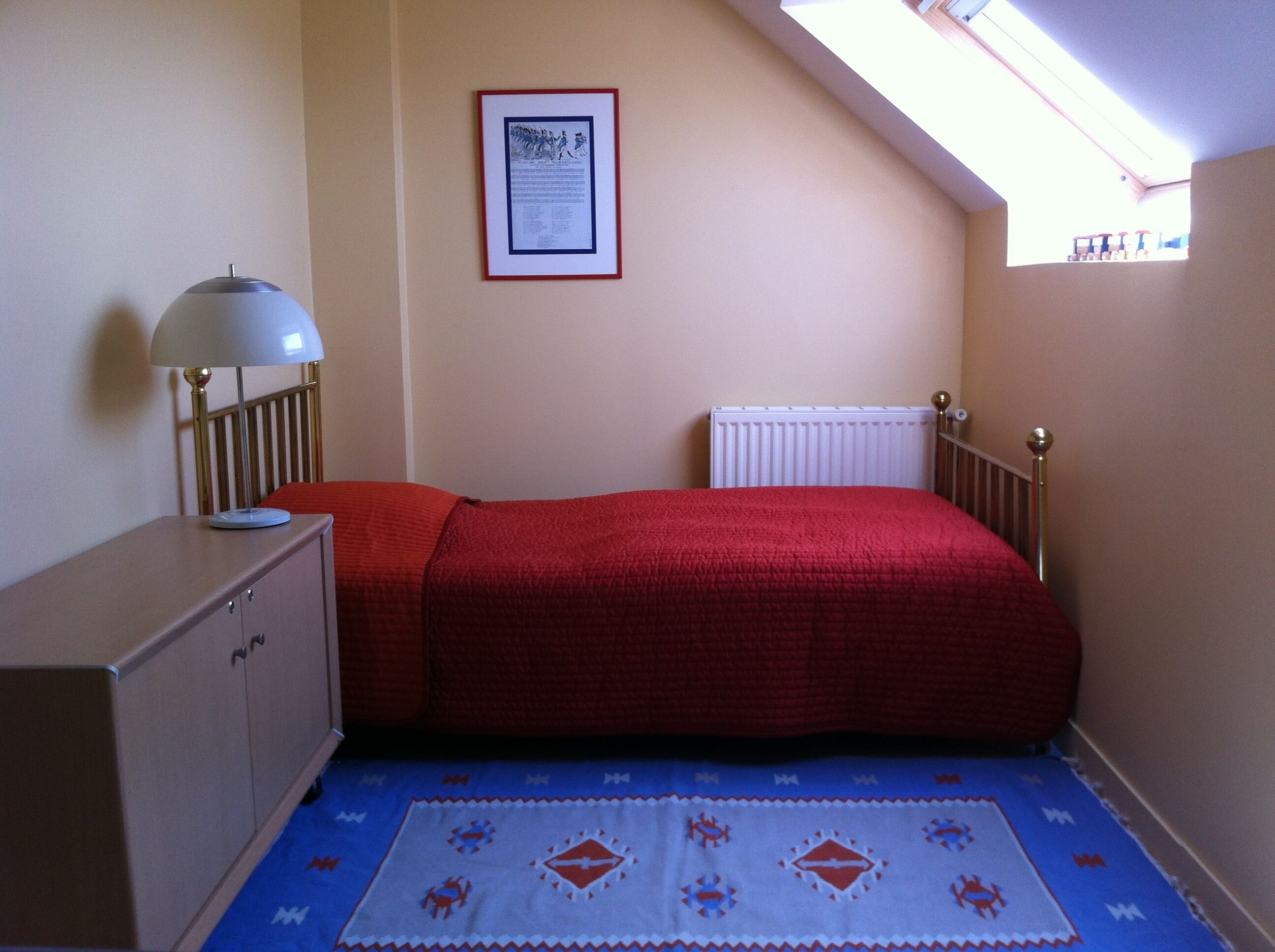 A small, single-bed room with a sloped ceiling, a radiator, a bedside table with a lamp, and a framed certificate on the wall.