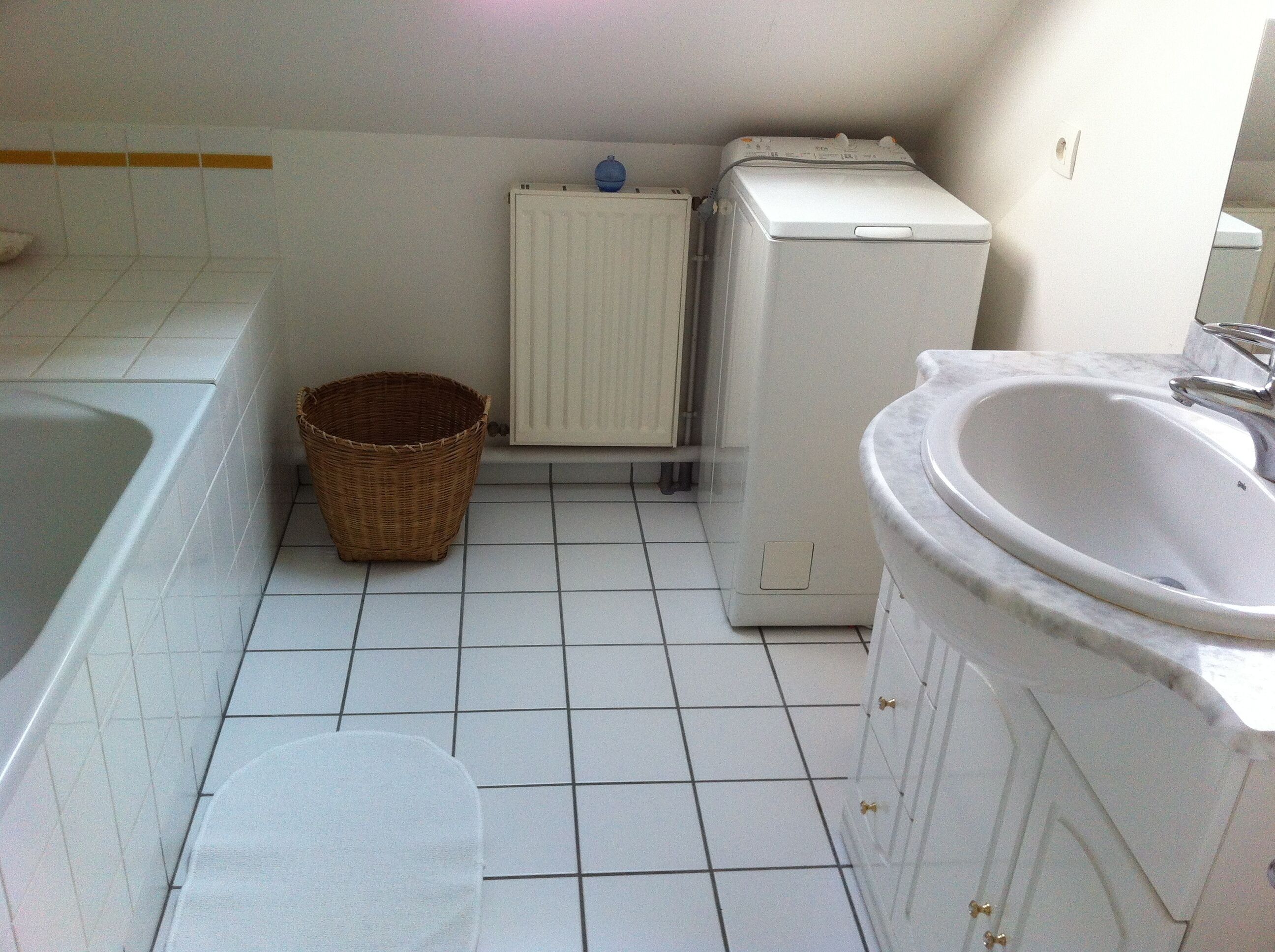 Une petite salle de bain carrelée, avec un lavabo blanc, des toilettes blanches, un radiateur blanc et un panier en osier.