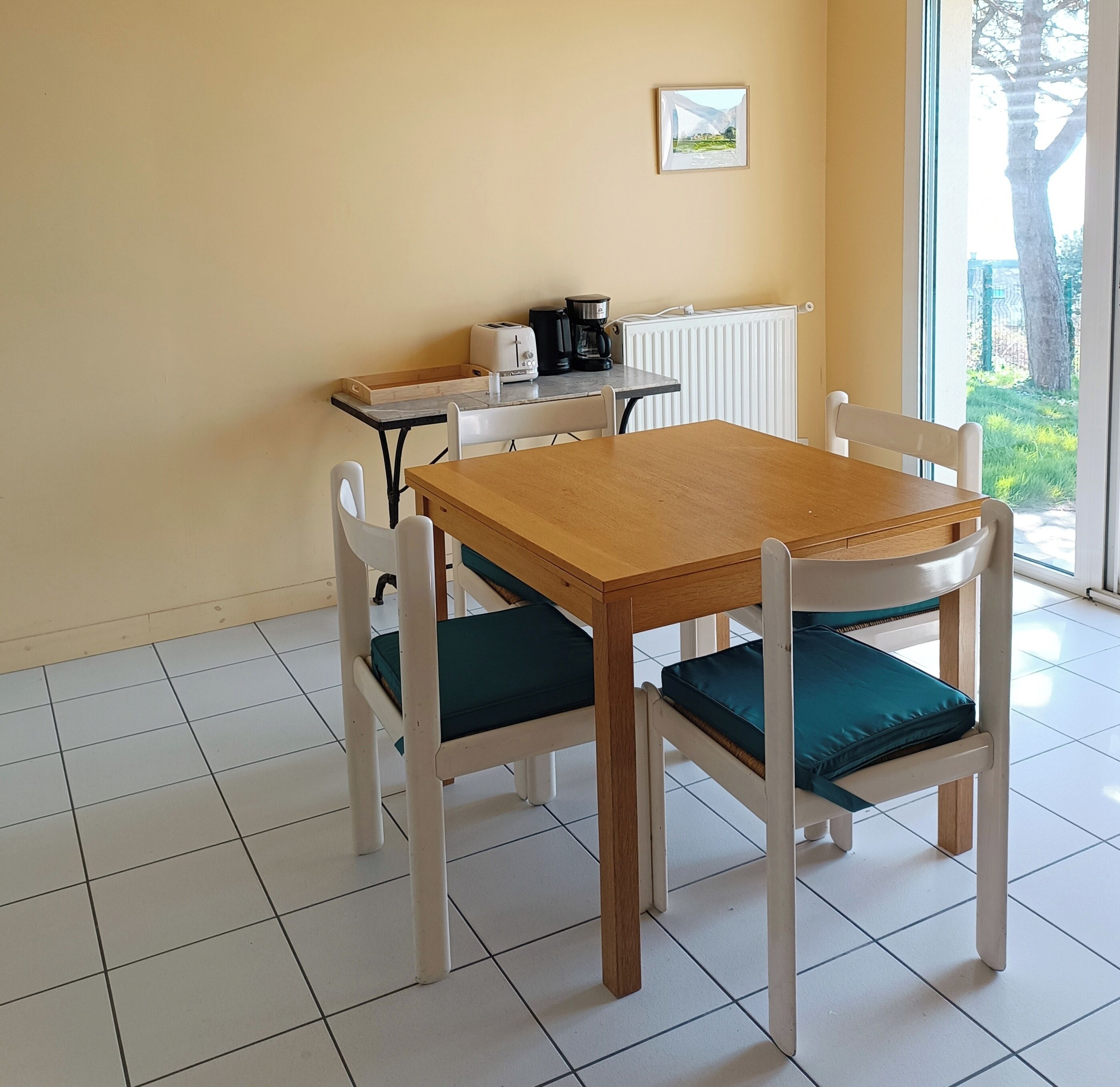 Een eethoek met een houten tafel en witte stoelen met teal kussens. Op de tafel staan een koffiezetapparaat en een broodrooster.