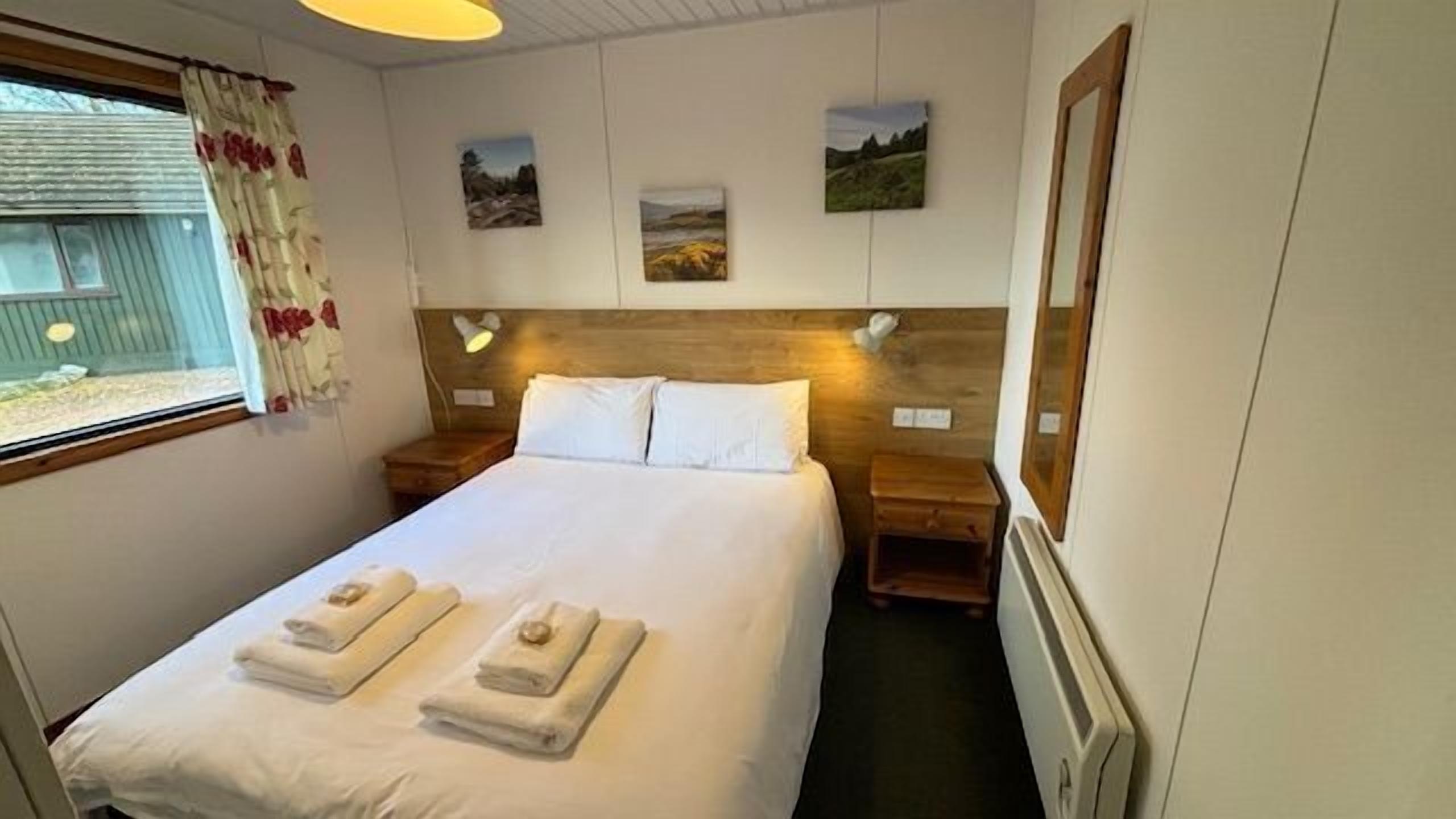 A bedroom with a bed, two bedside tables, a window with curtains, and three wall-mounted pictures.