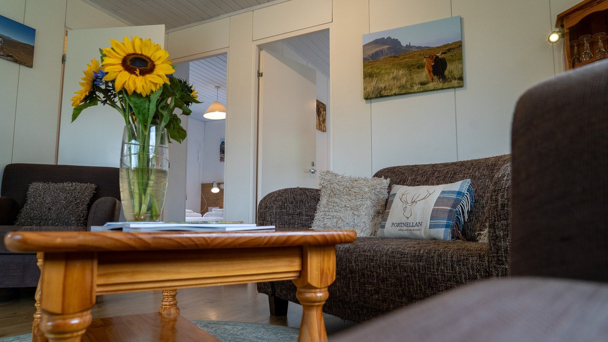 A living room with a sofa, a coffee table, and a vase with sunflowers. There are paintings on the walls and a door leading to another room.