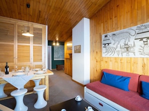 A cozy room with a wooden ceiling, a dining area with a table set for four, and a red sofa with blue cushions.