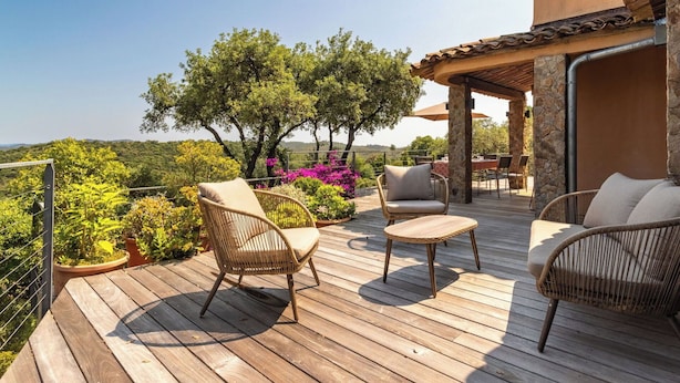 Une terrasse en bois avec des meubles en osier, un pilier en pierre et une vue sur la verdure et les collines.