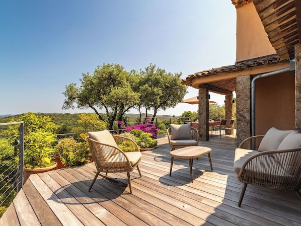 A wooden deck with wicker furniture, a stone pillar, and a view of greenery and hills.