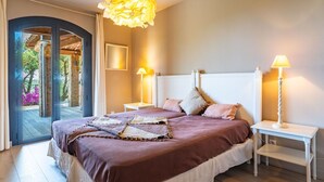 Une chambre avec un grand lit, deux tables de chevet, un luminaire suspendu et une vue sur l’extérieur, visible à travers une porte en arc de cercle.
