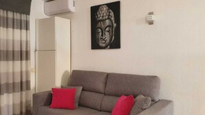 Una sala de estar con un sofá gris, cojines rojos, una estatua de Buda y una ventana con cortinas a rayas.