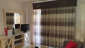Una sala de estar con un televisor sobre un mueble blanco, una mesa de vidrio y una cortina a rayas.