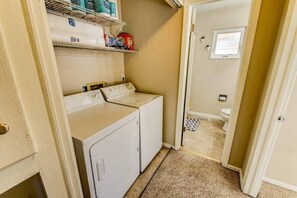 Ein Waschbereich mit Waschmaschine und Trockner, ein Regal mit Reinigungsmitteln und Blick in ein Badezimmer mit Toilette und Fenster.