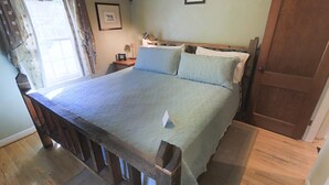 Une chambre avec un lit en bois, une fenêtre avec des rideaux, une table de chevet avec une lampe et un tableau encadré au mur.