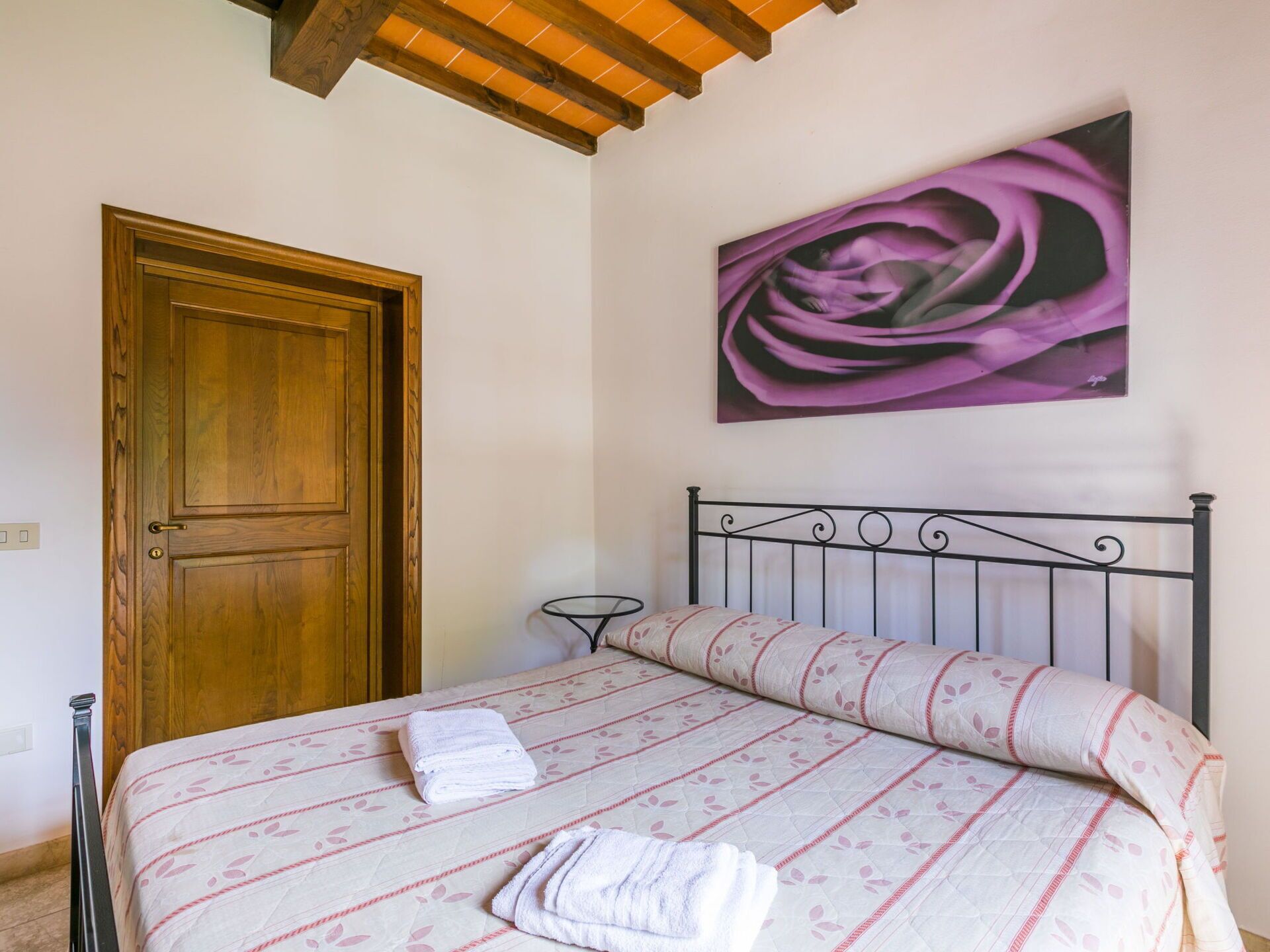 A bedroom with a wooden door, a bed with a patterned bedspread, a metal headboard, and a painting of a purple flower on the wall.