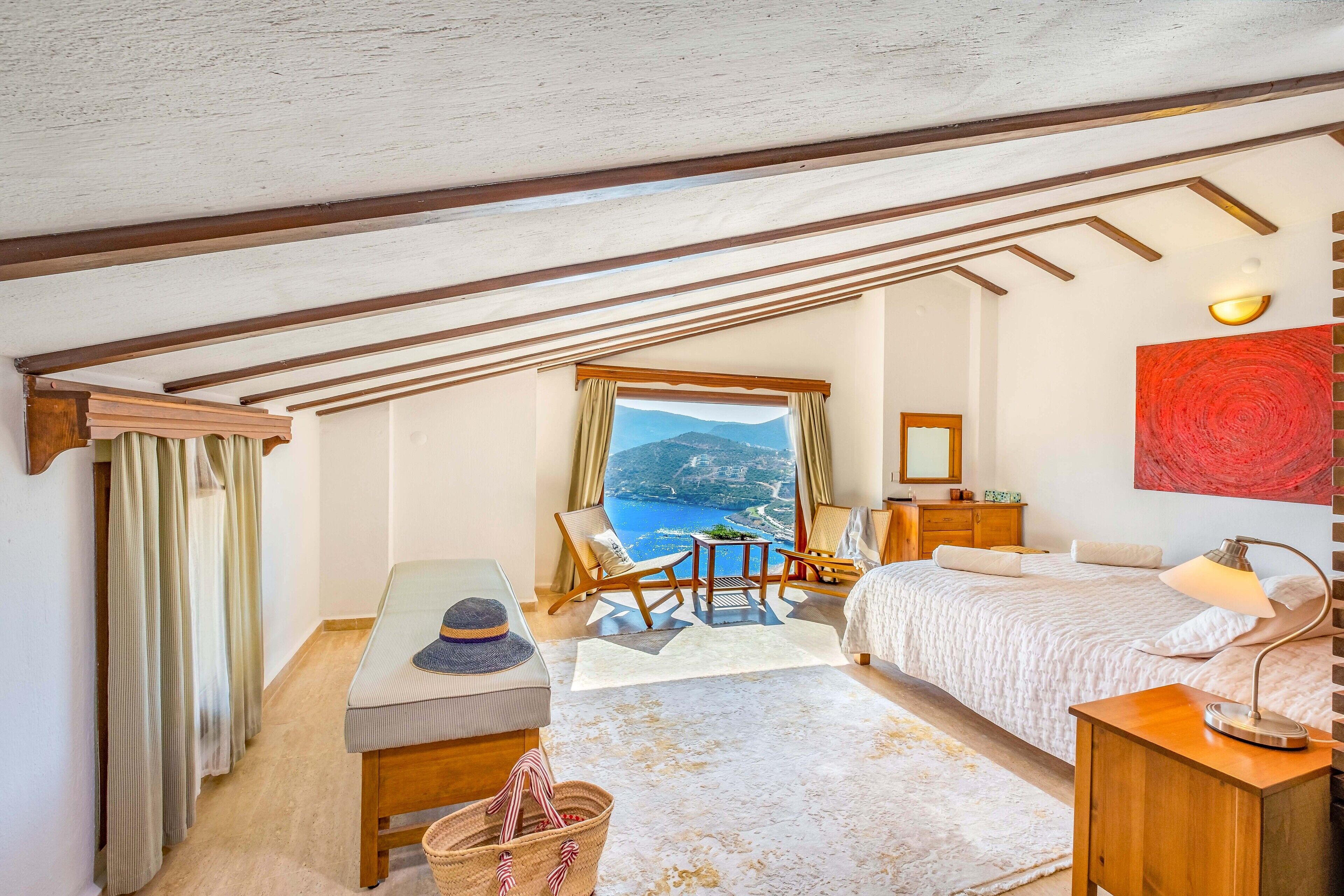Une chambre à coucher comprenant un lit, un bureau, une chaise, une fenêtre donnant sur la mer et un tableau abstrait rouge accroché au mur.