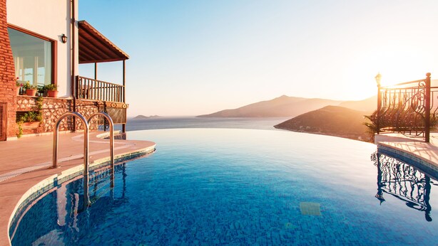A infinity pool with clear blue water, a wooden deck, and a stone wall. - Secluded & Luxurious Mediterranean Villa w/ Heated Infinity Pool & Kids' Pool (Kalkan)