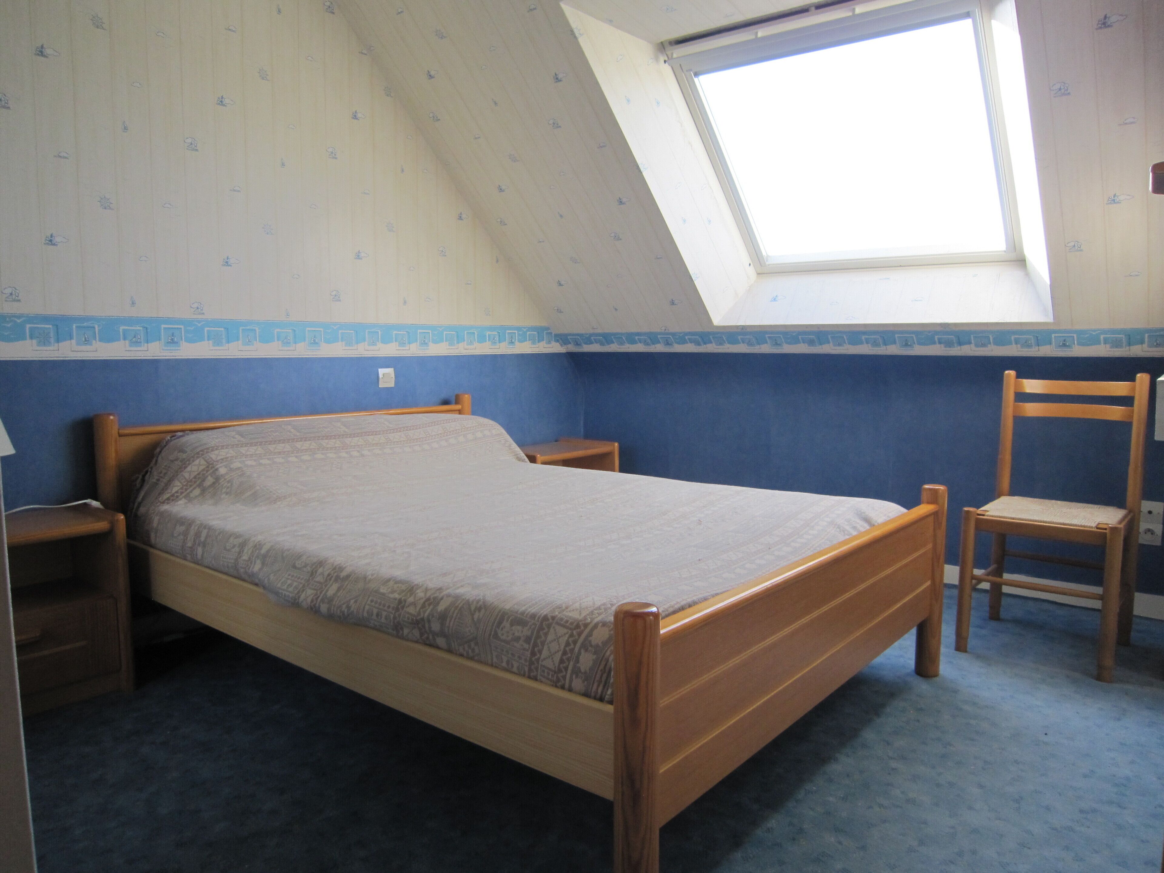 Un dormitorio con techo inclinado, tragaluz, cama, silla y mesita de noche.