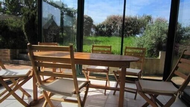A dining area with a wooden table and chairs, large windows offering a view of greenery outside.