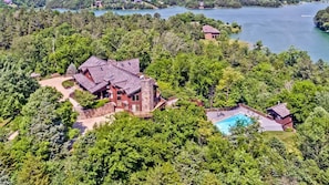 A large house with a stone chimney, surrounded by dense trees and a lake.