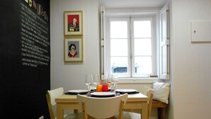 Un comedor con una mesa dispuesta para dos, dos sillas, una ventana con contraventanas blancas y una pared decorada con cuadros.