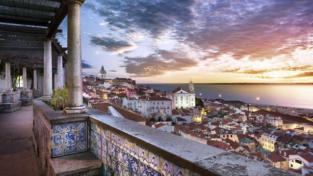 Un paisaje urbano costero al atardecer, con edificios históricos y una estructura con cúpula a lo lejos.