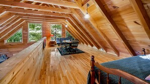 Ein Schlafzimmer im Holzbau mit geneigter Decke, einem Bett, einem Esstisch mit Stühlen und Blick auf Bäume durch große Fenster.