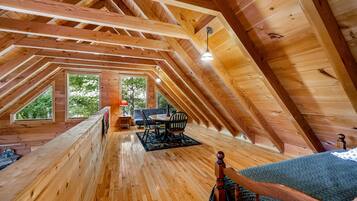 Une chambre mansardée en bois, avec un plafond en pente, un lit, une table à manger avec des chaises et une vue sur les arbres à travers de grandes fenêtres.